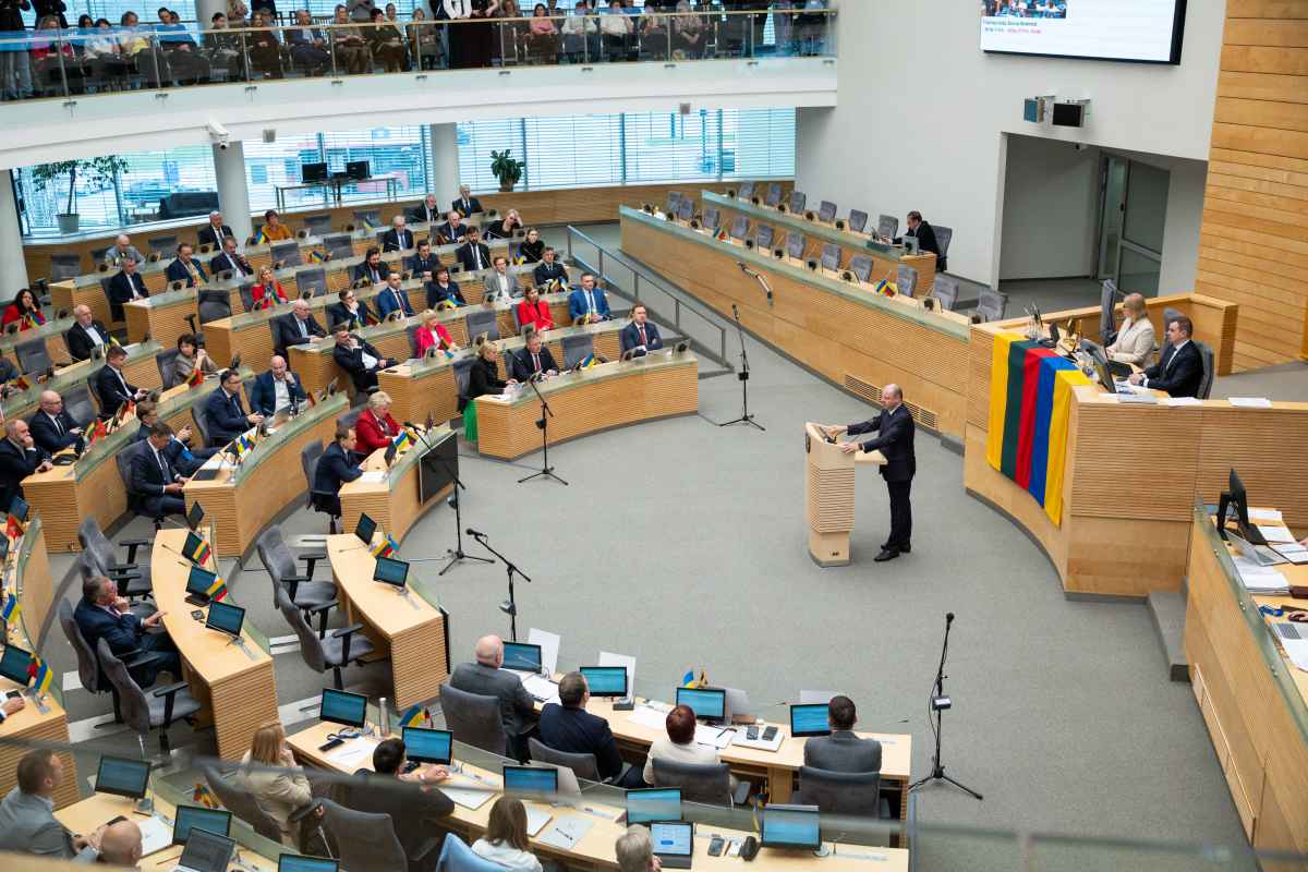Žmonės sėdi parlamento salėje klausosi kalbančio vyro prie pulto